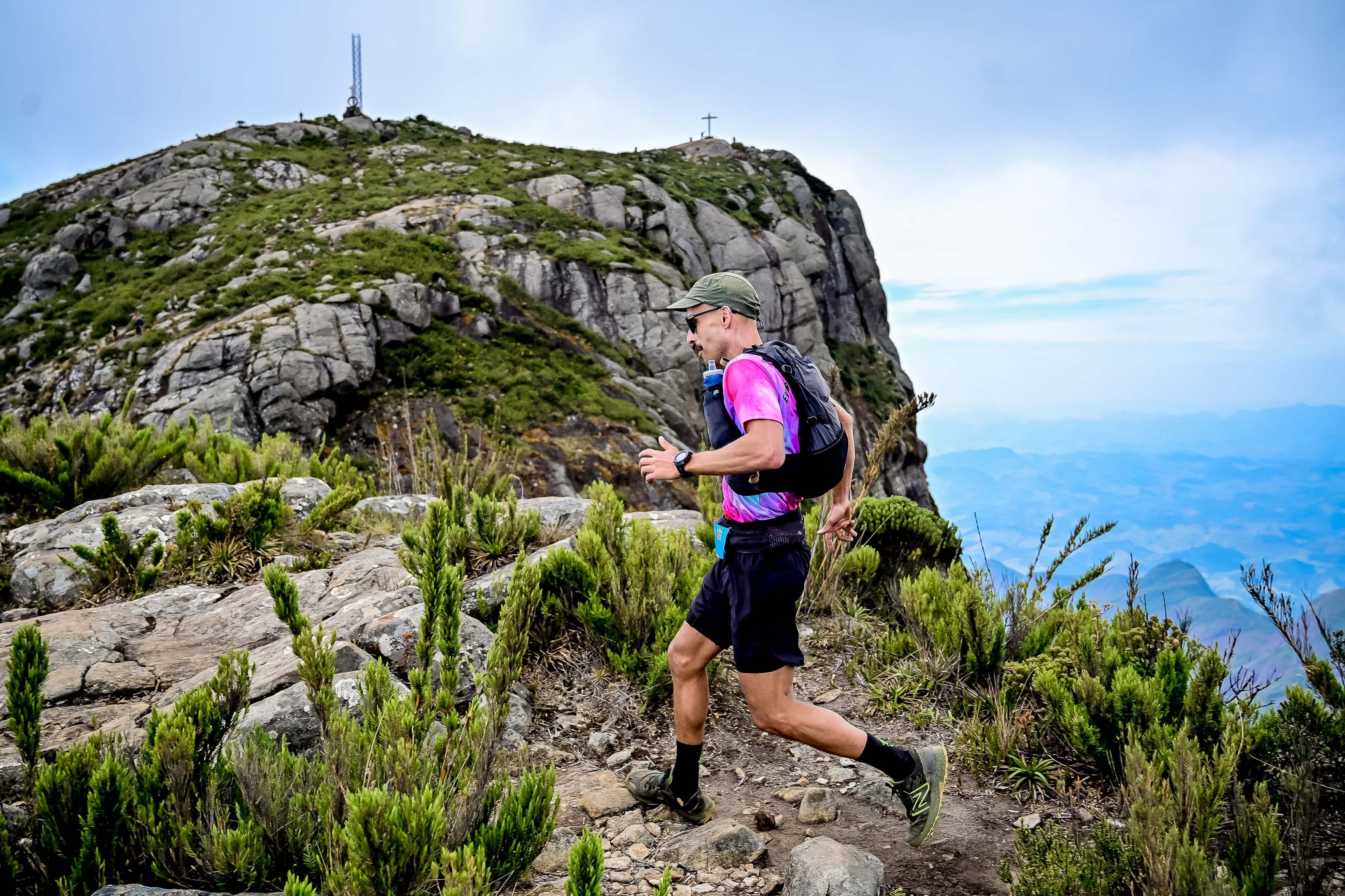 Corredor de trilha subindo o cume rochoso do Pico da Bandeira, com o vale azulado se abrindo à direita