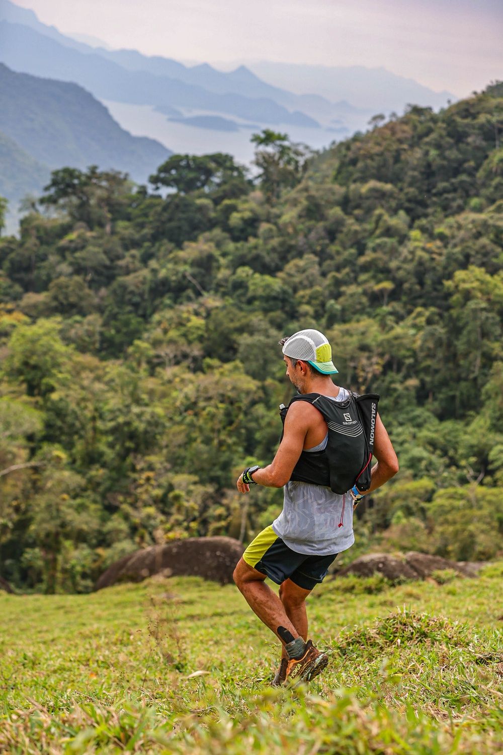 Corredor de costas descendo um campo de capim, com mata fechada, serras e o mar de Paraty ao fundo